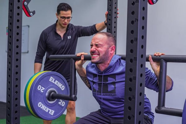 Muscular sportsman in activewear squatting with heavy barbell with effort while listening trainer in gym