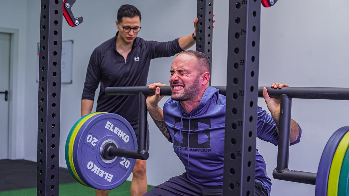 Muscular sportsman in activewear squatting with heavy barbell with effort while listening trainer in gym